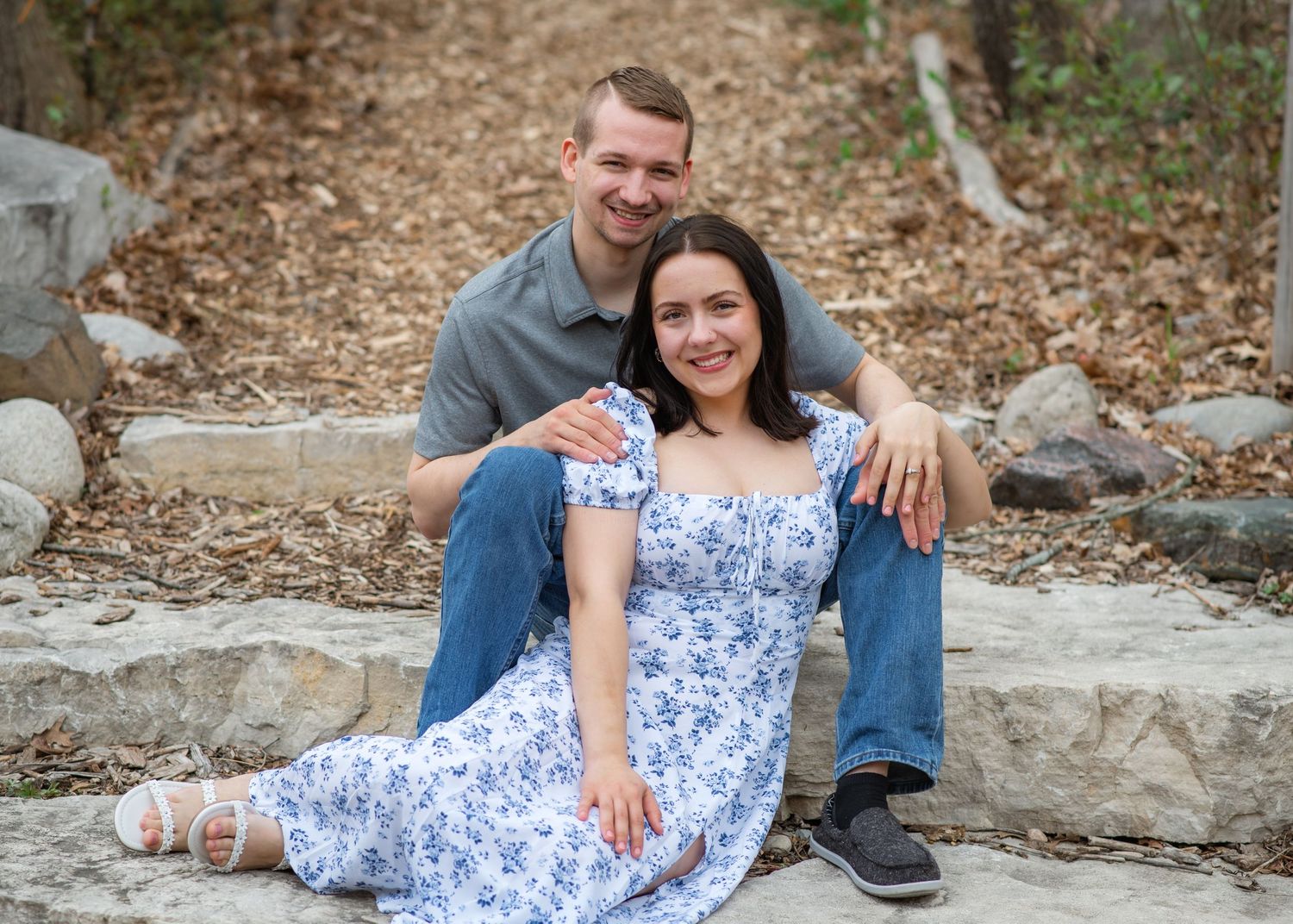 A couple sits together on stone steps outdoors, with person in a blue floral dress and jeans visible in the scene.