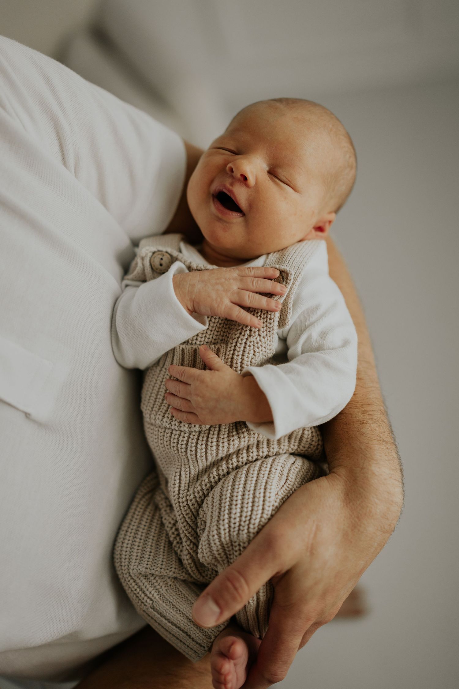 A baby wearing a beige knit romper is held gently while laying on white bedding.
