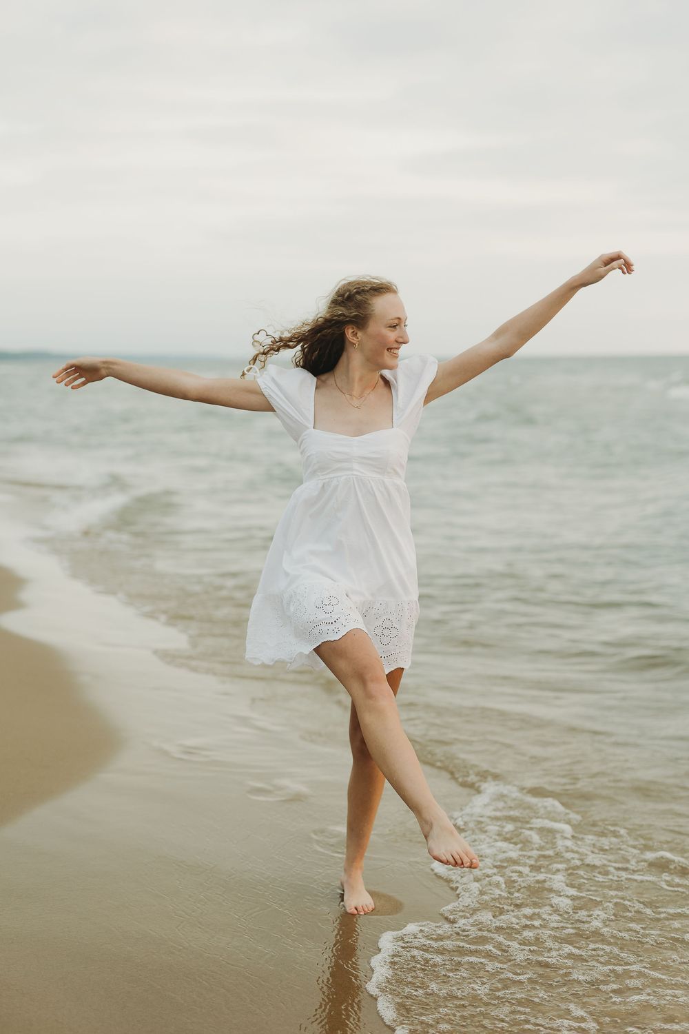 A dancer in white dress twirls gracefully along the shoreline at sunset with ocean waves gently rolling in.