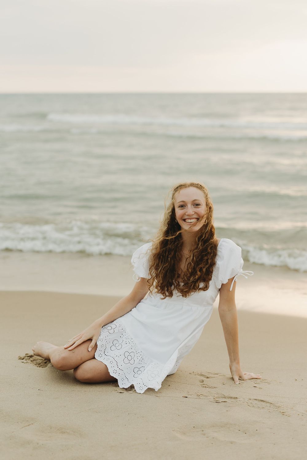 A woman in a white lace dress lying gracefully on a sandy beach during sunset.