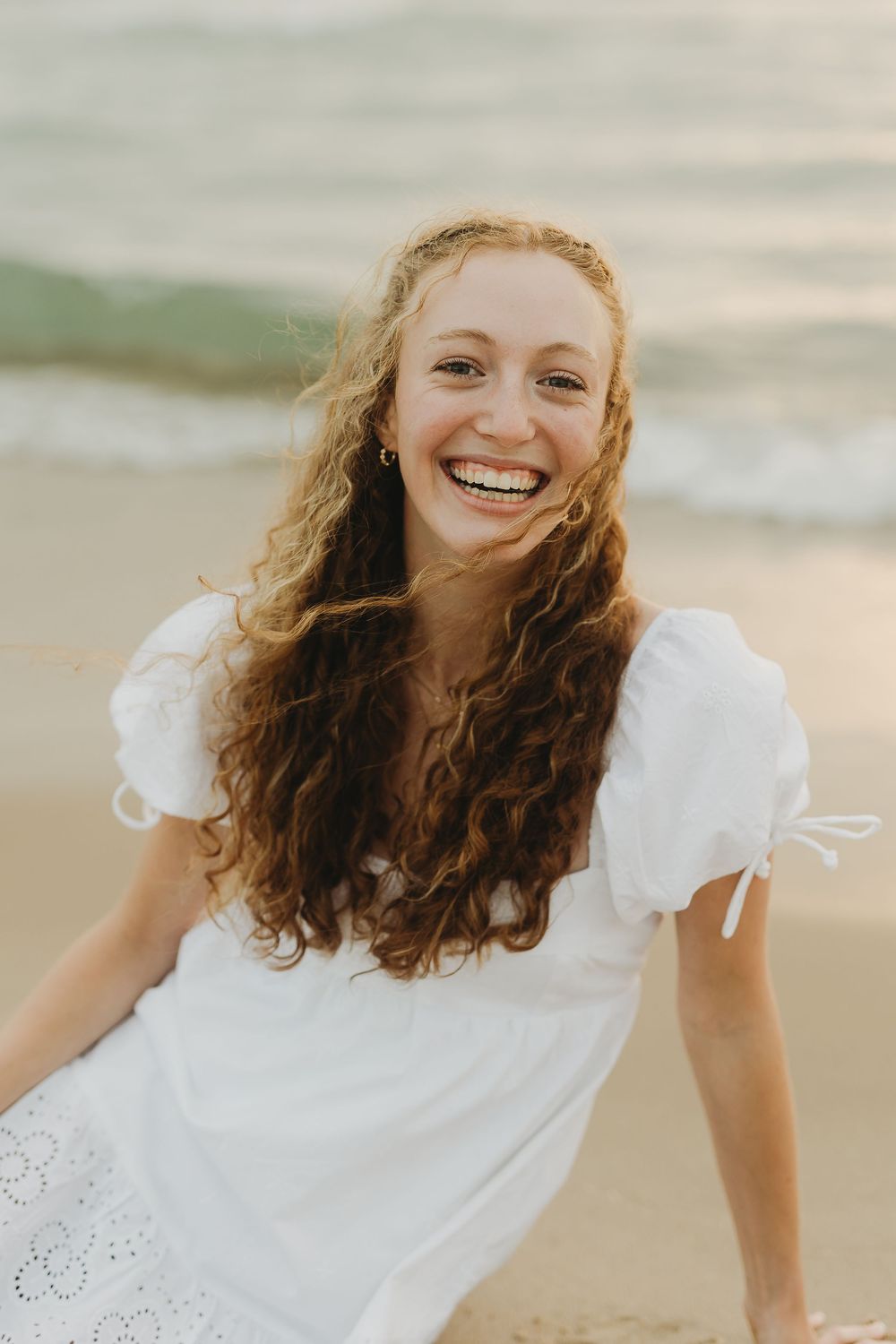 Woman in white flowing dress with curly hair smiles joyfully on a sunny beach.