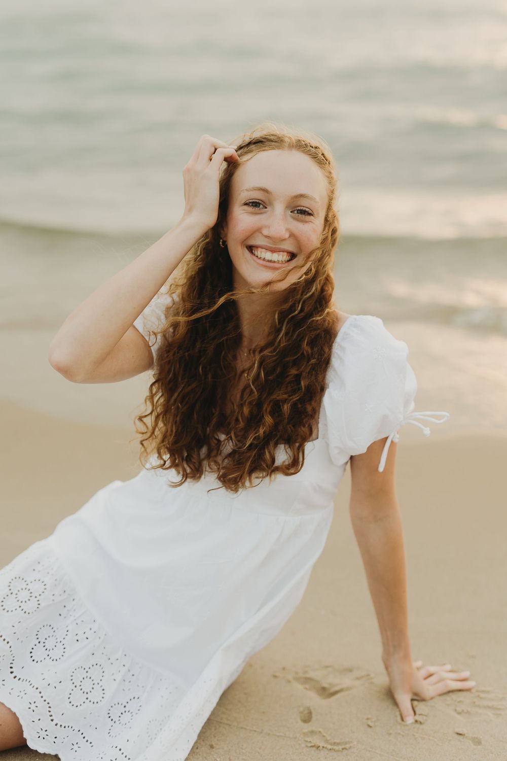 A person with long curly hair wearing a white eyelet dress smiles while sitting on a beach at sunset.