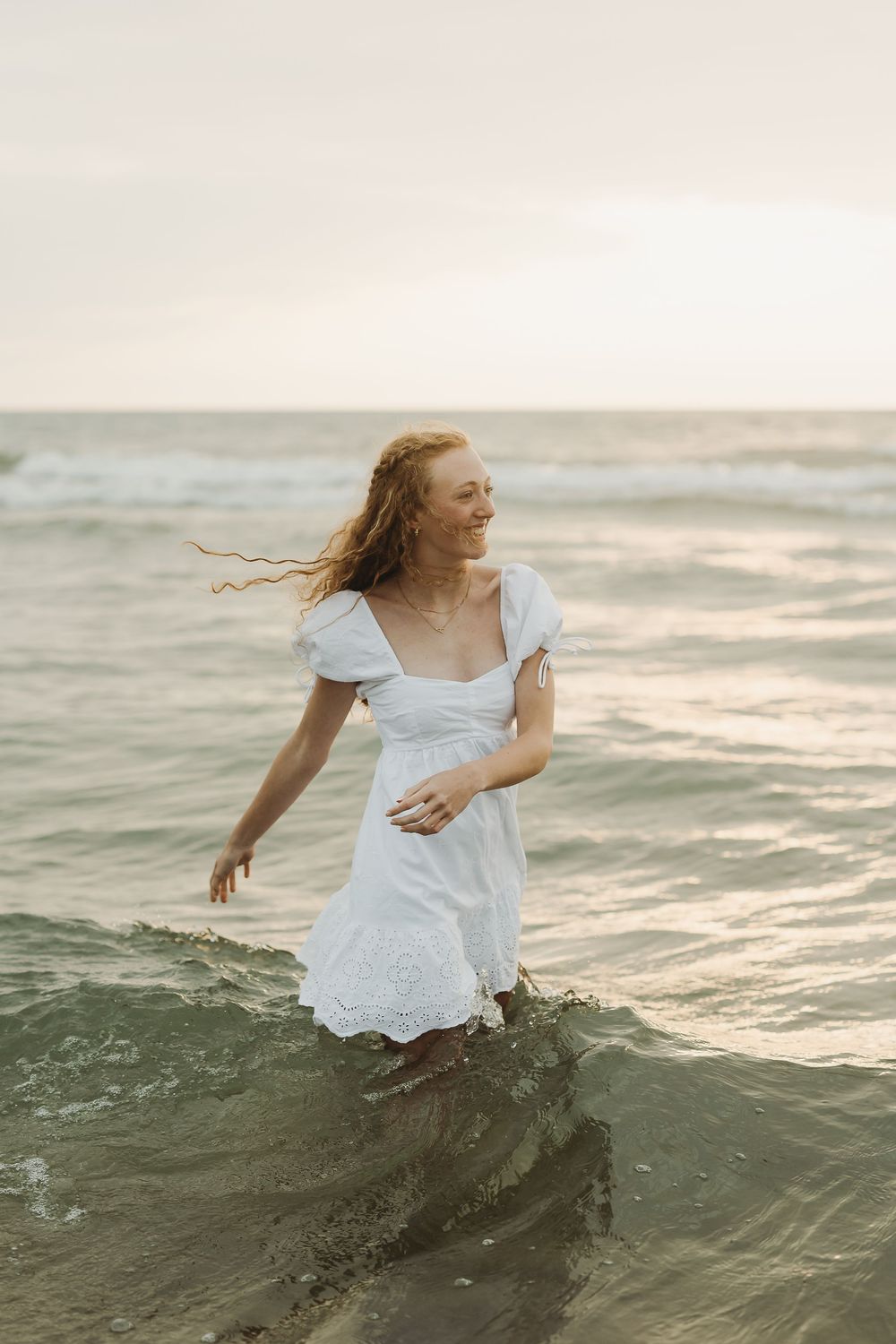 A woman in a white dress dances gracefully in the ocean waves during sunset, creating a dreamy seaside scene.