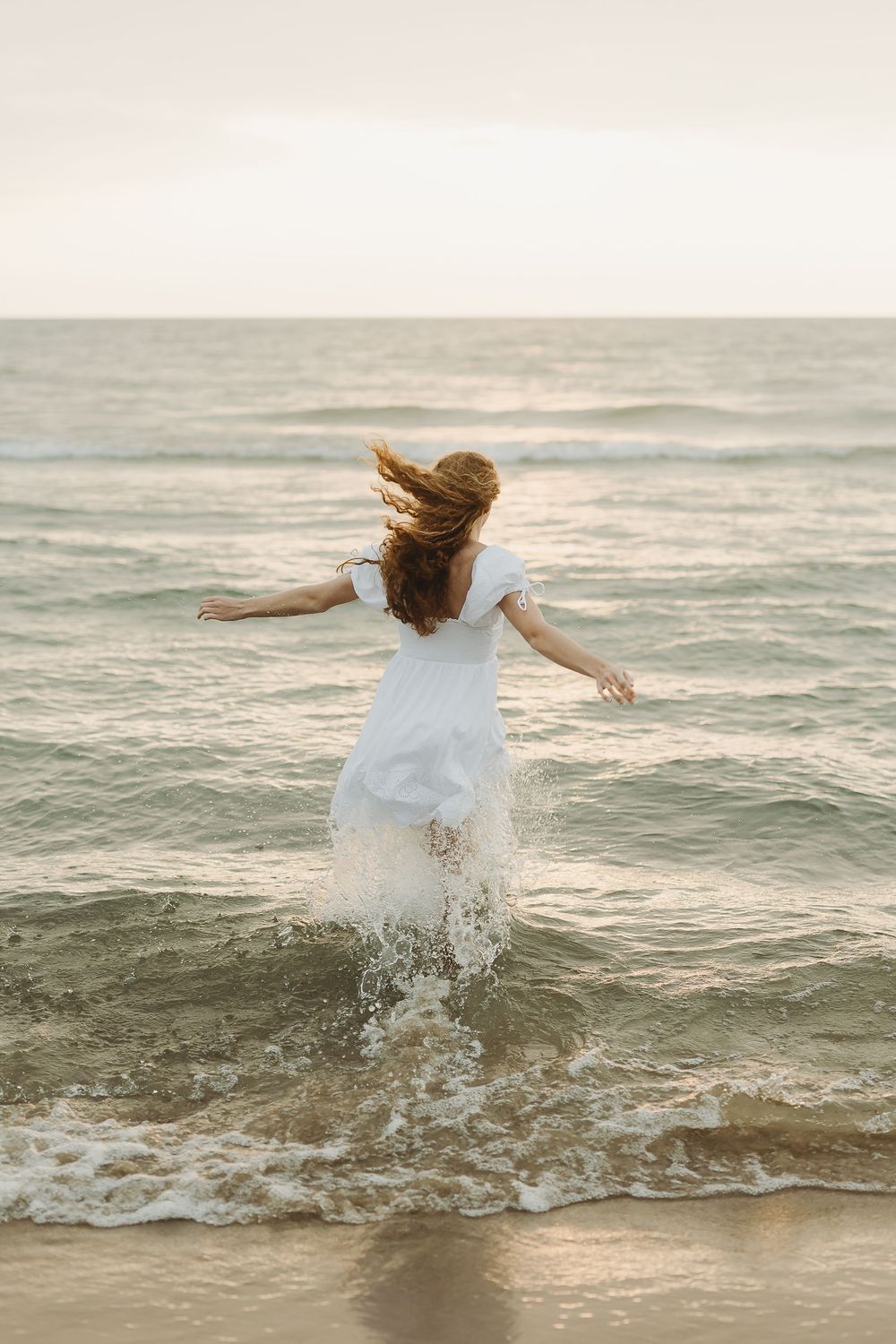 A person in a flowy white dress dances and splashes through ocean waves at sunset, creating a dreamy beach scene.