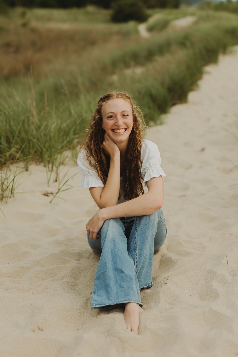 A smiling person in casual denim and white shirt sits barefoot on a sandy path with beach grass on a sunny day.