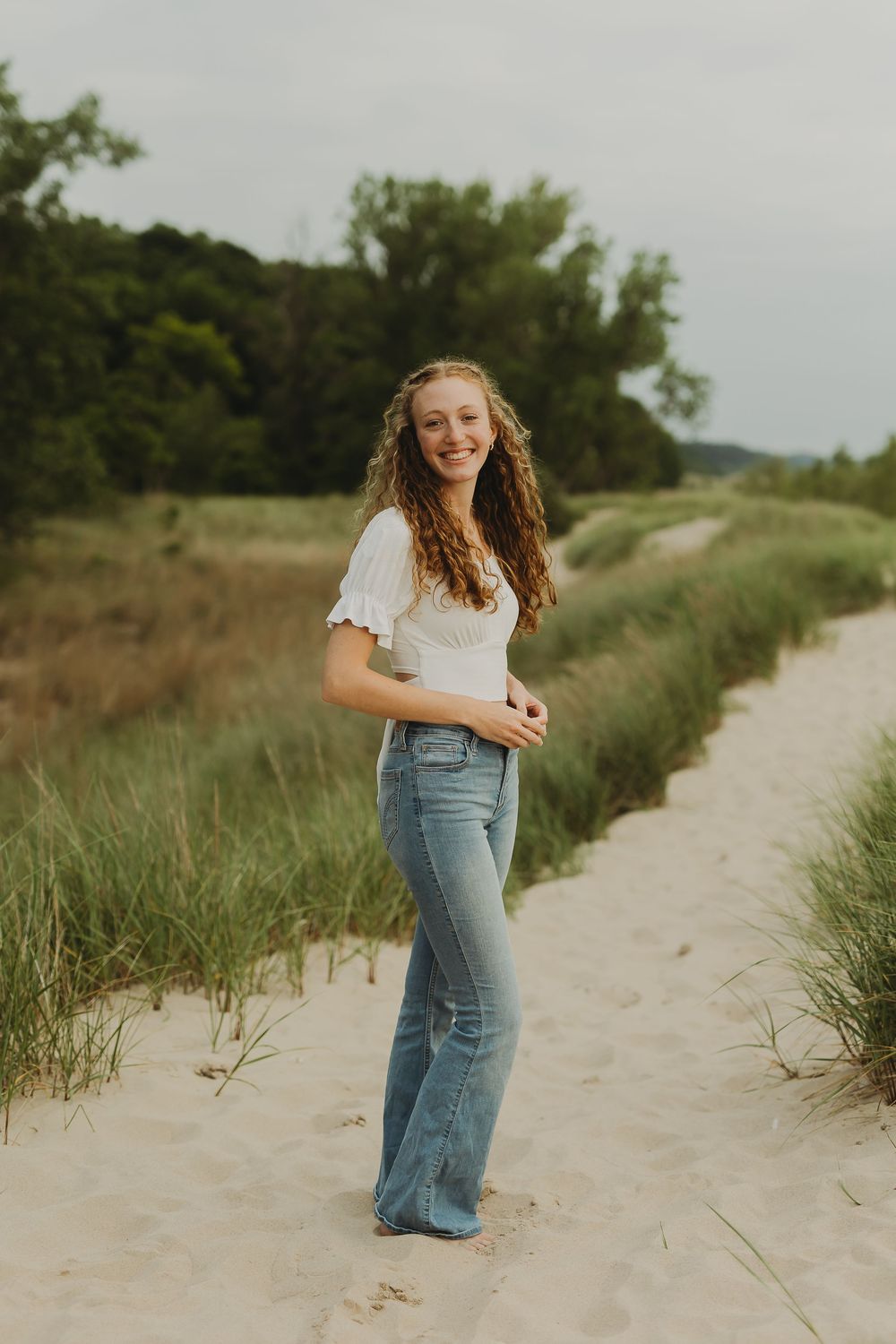 A person in jeans and white top walks along a sandy beach path surrounded by tall grasses on a summer day.