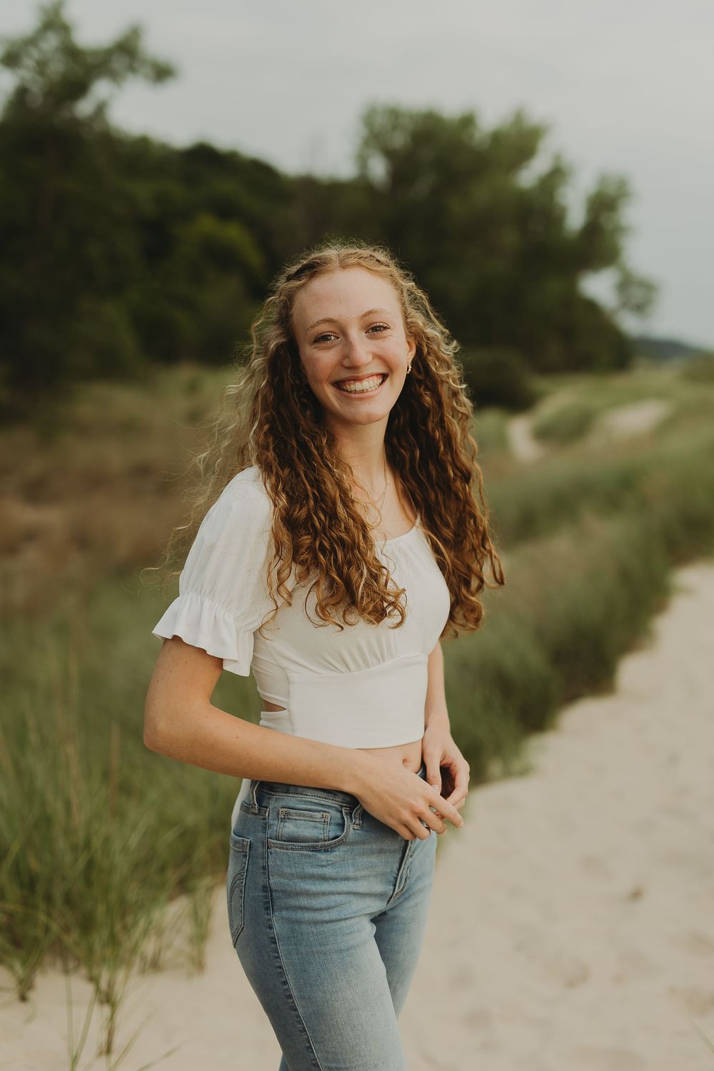A person with long wavy brown hair smiles in a white crop top and jeans while standing on a sandy path.