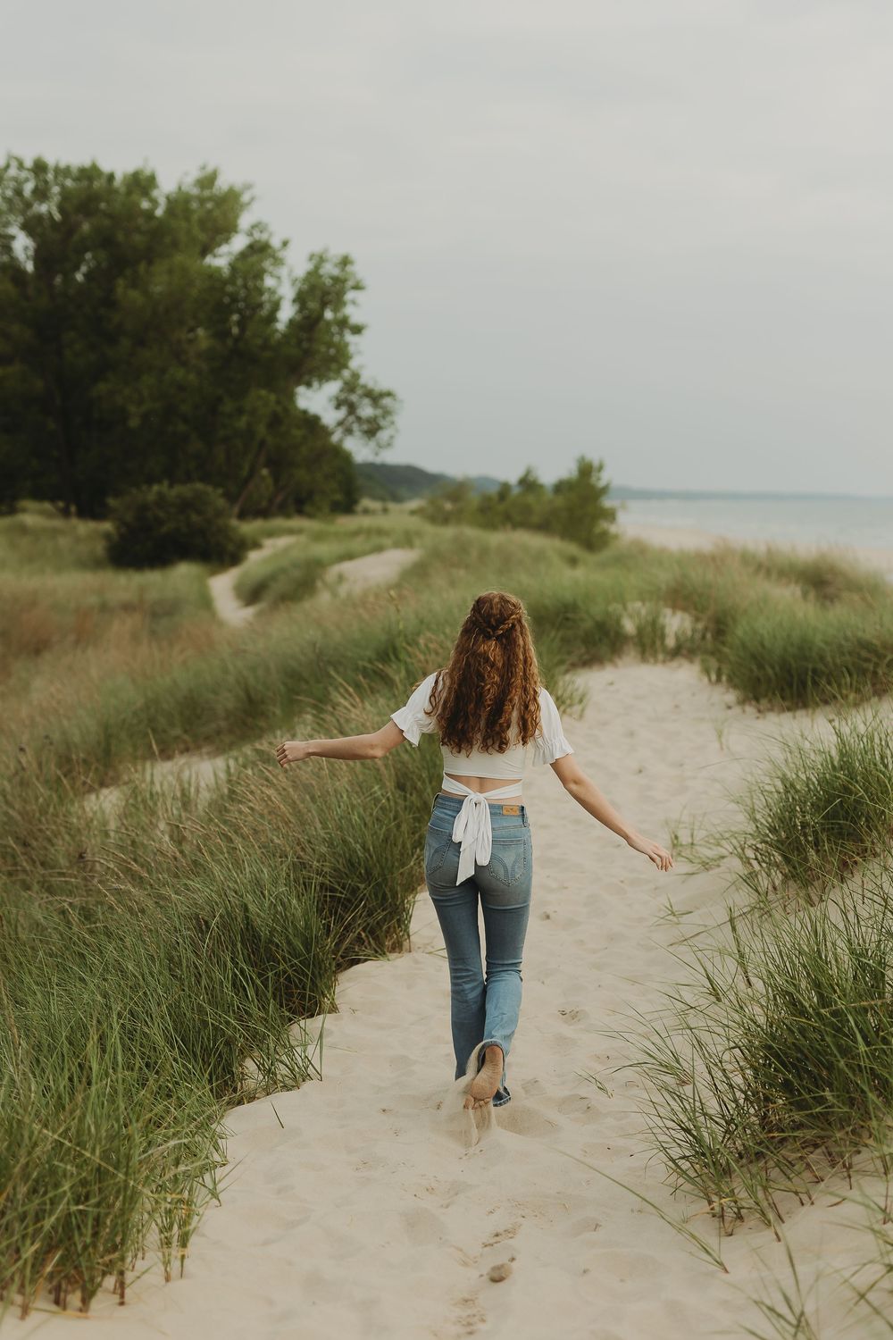 A wanderer in jeans walks along a sandy beach path bordered by tall grass on a cloudy summer day.