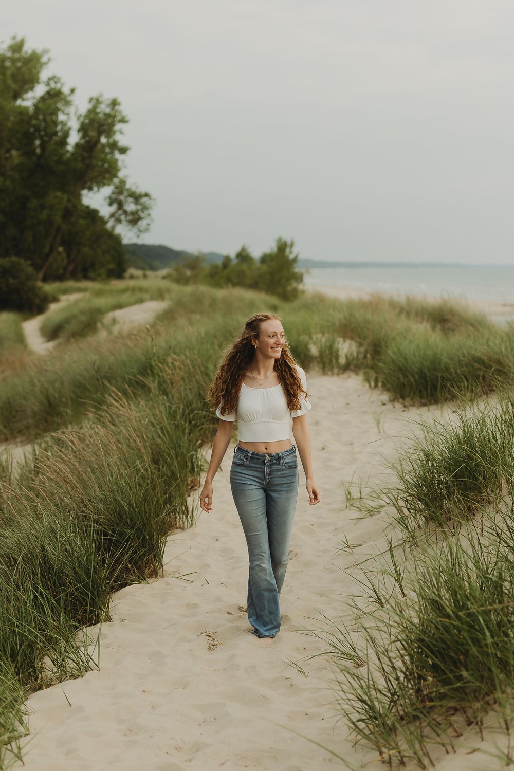 Woman in jeans walks along a sandy beach path bordered by tall dune grass on a sunny summer day near the water.