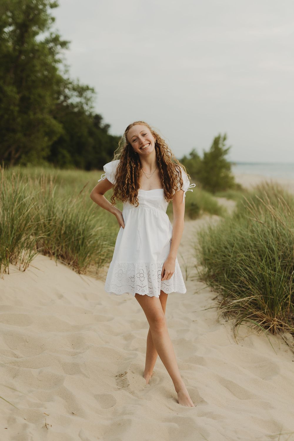 A carefree dancer in a white dress twirls on a sandy beach dune with tall grass and trees in the background.