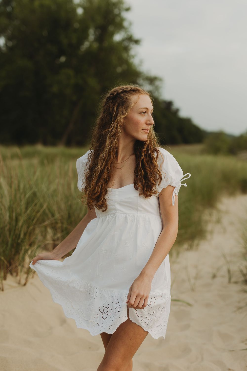 A figure in a flowing white dress dances and twirls on a sandy beach with natural grass in the background.