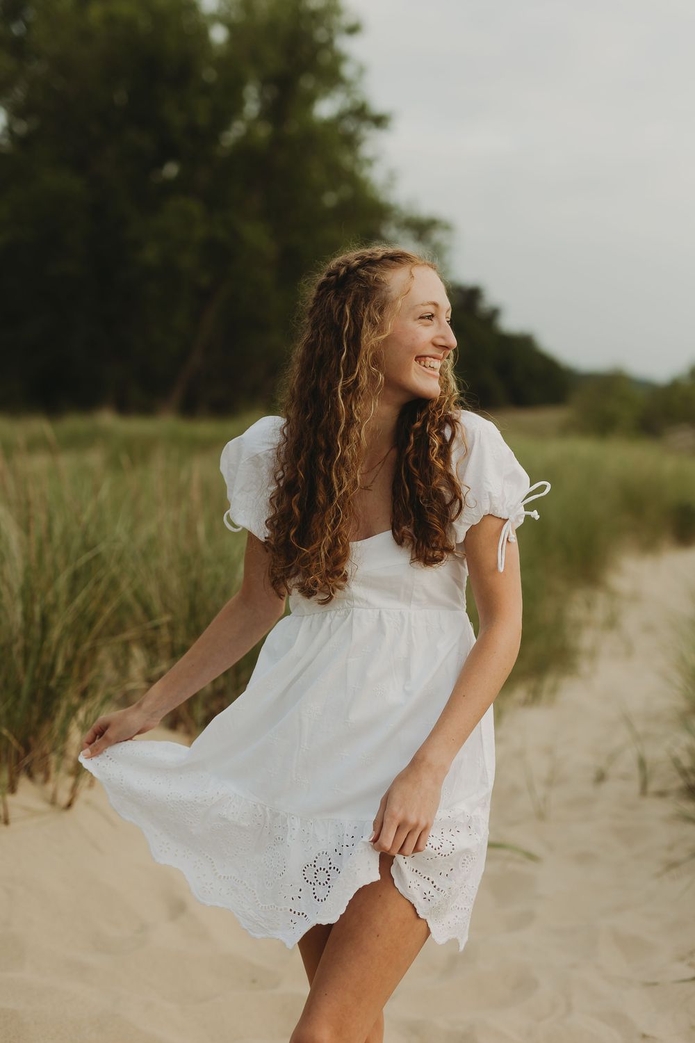 A carefree figure in a white dress twirls playfully on a sandy beach path surrounded by greenery.
