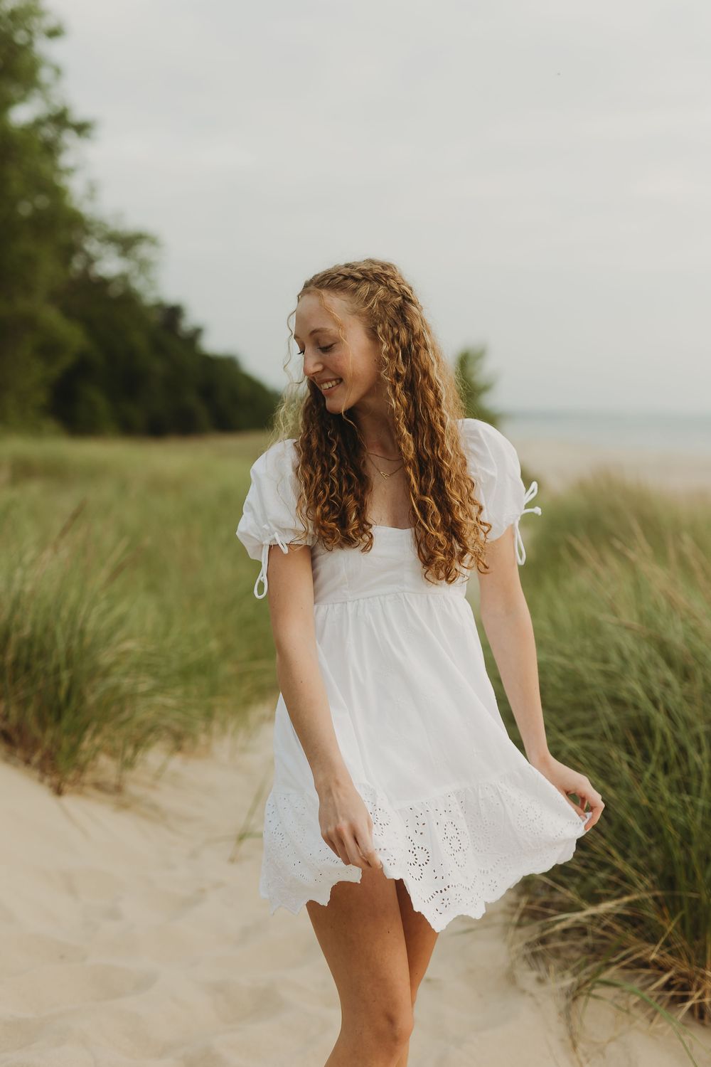A figure in a flowing white lace dress stands poetically among beach grass and sand dunes on a summer day.