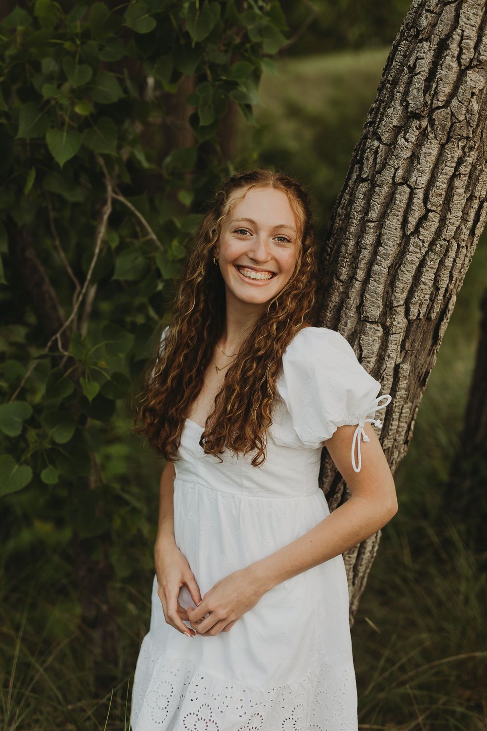 A figure in a white eyelet dress leans against a tree trunk in a natural outdoor setting with green foliage.