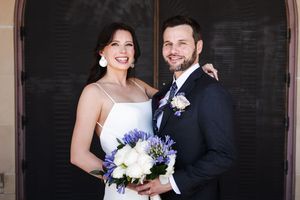 A joyful wedding portrait in front of an arched doorway with purple and white bouquet.