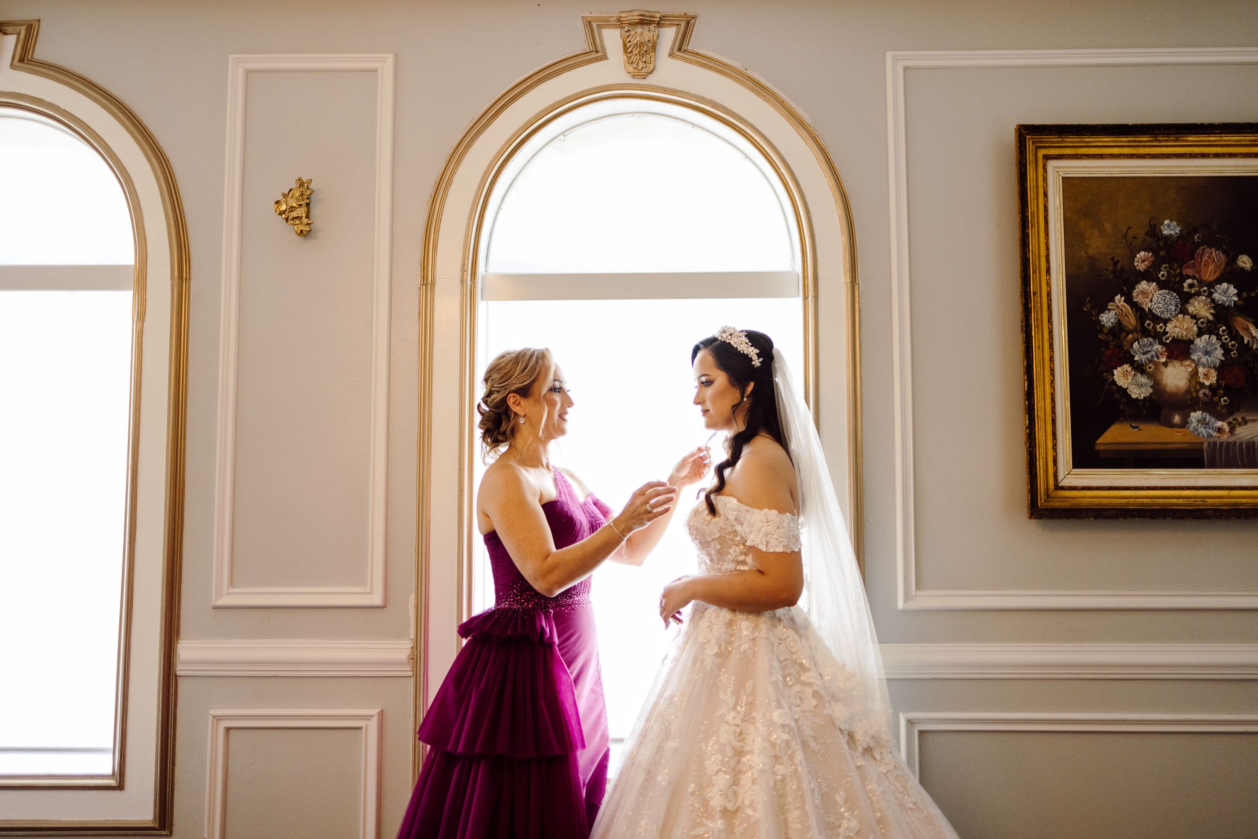 Two people in formal dresses share an intimate moment in an elegant room with arched windows and floral artwork.