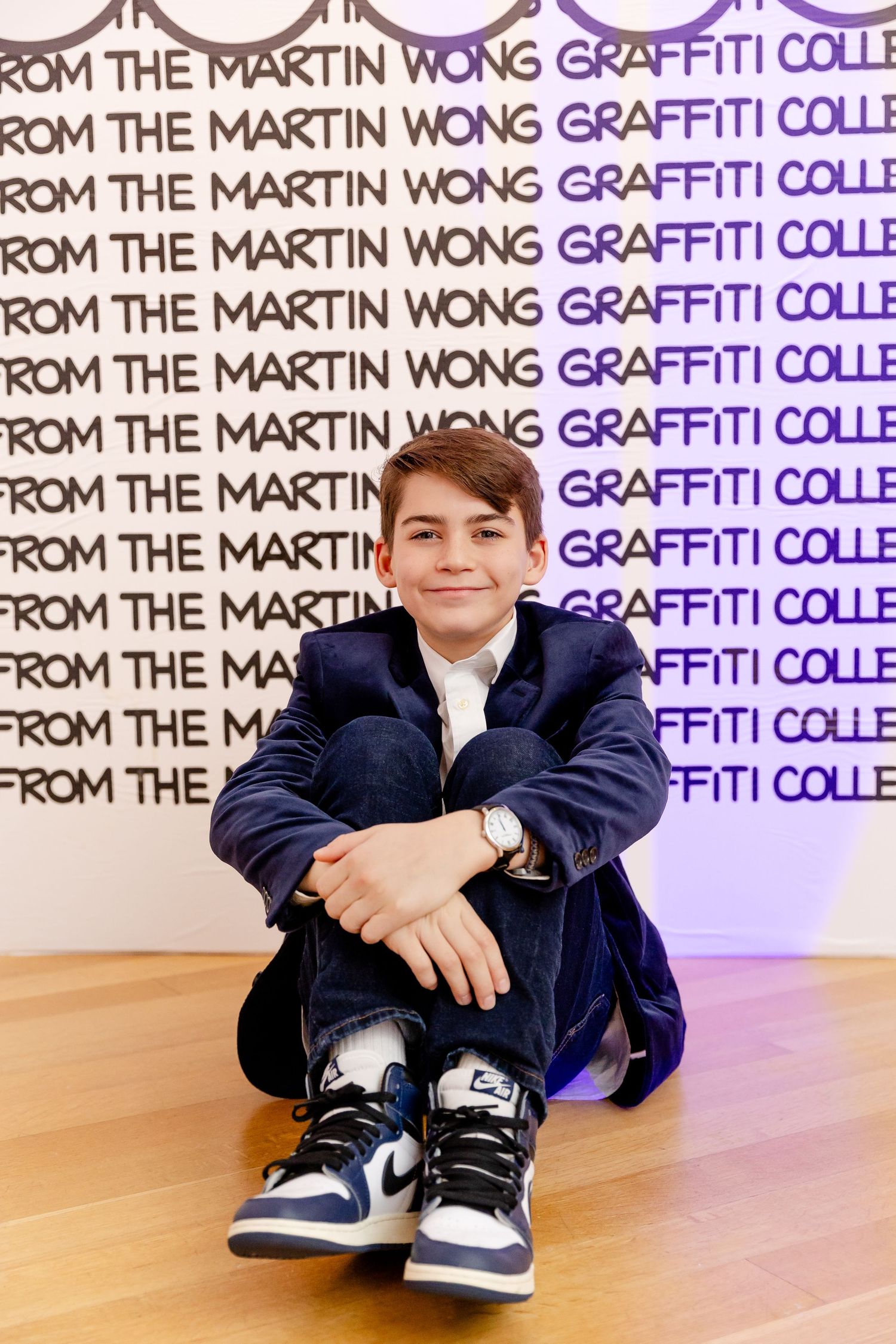 A figure sits crouched against a purple and white wall with repeating text about Martin Wong Graffiti Collection.