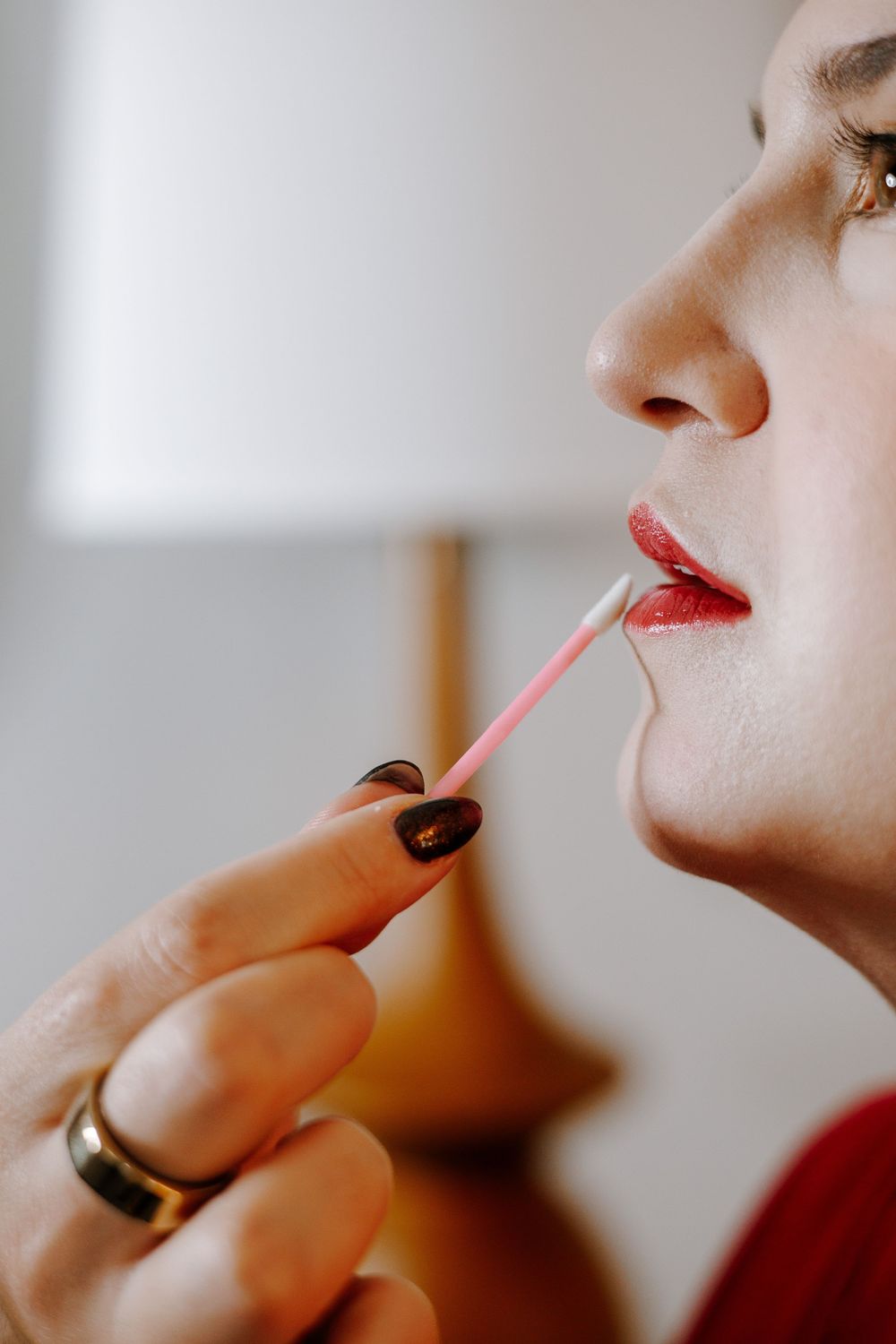 Close-up of a woman applying red lip gloss with a wand, wearing a gold ring and dark nail polish.