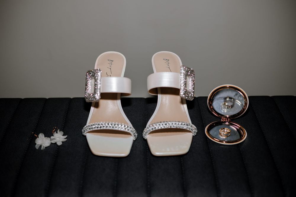 White bridal heels with crystal embellishments displayed beside floral earrings and ring compact mirror.