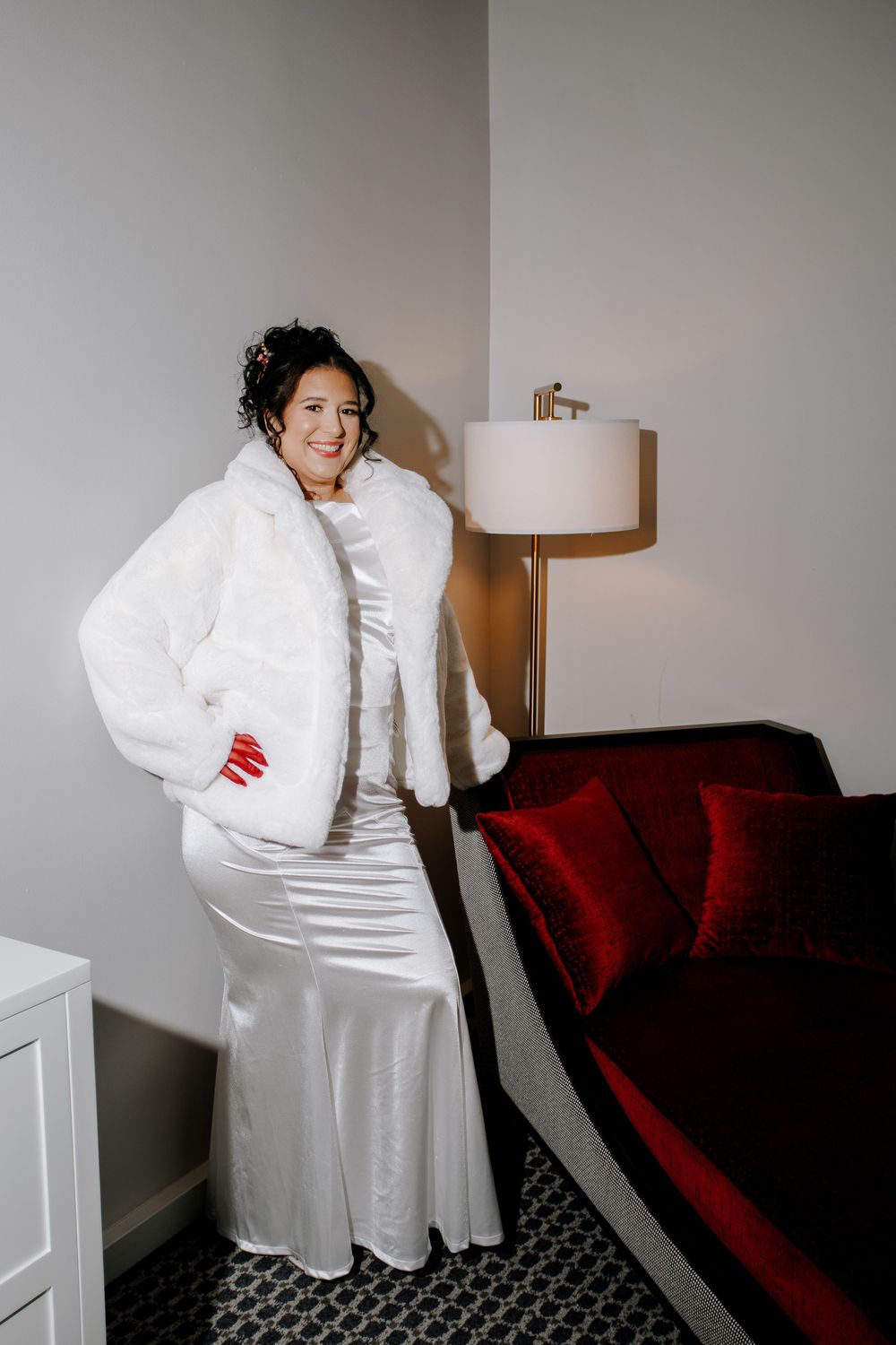 Radiant bride poses in a white satin gown and luxurious white faux fur jacket in a hotel room.