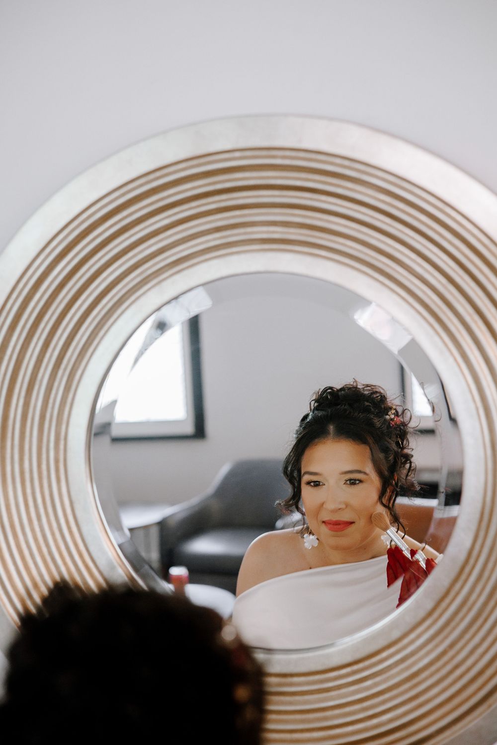 Beautiful bride with curly updo and red lips gets makeup applied, reflected in a round decorative mirror.