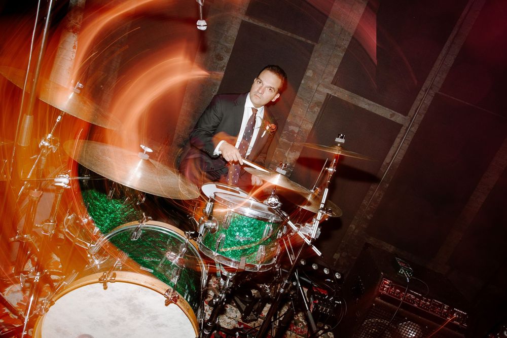 Man in suit playing green drum kit on stage with dynamic motion blur effect under warm orange lighting.
