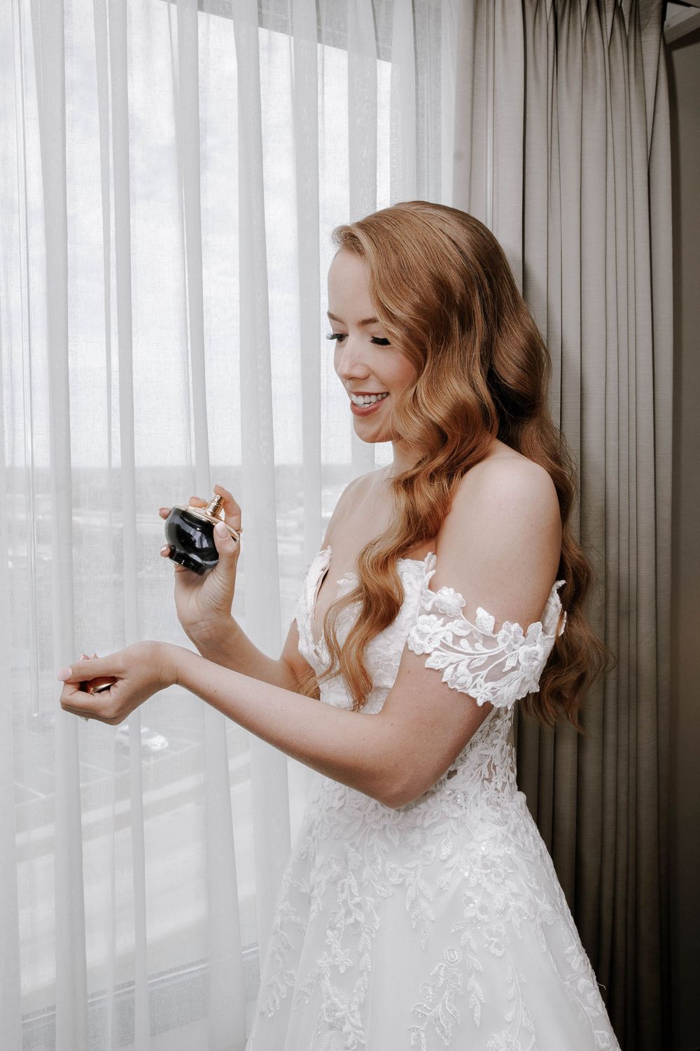 Smiling bride in lace off-shoulder wedding gown applying perfume by sheer curtained window.