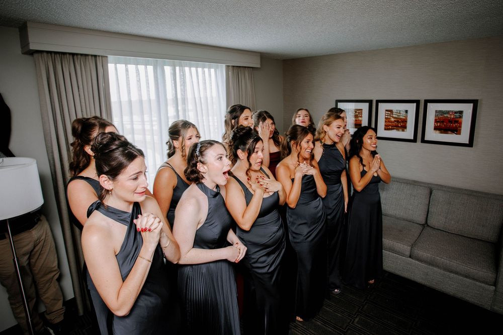 Bridesmaids in black gowns react with joy and emotion during a wedding morning getting-ready moment.