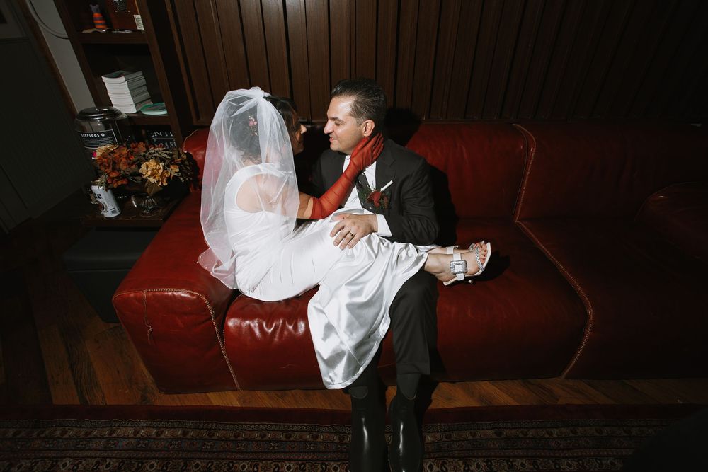 Groom in black suit with red scarf carries bride in white wedding dress and veil on a red sofa.
