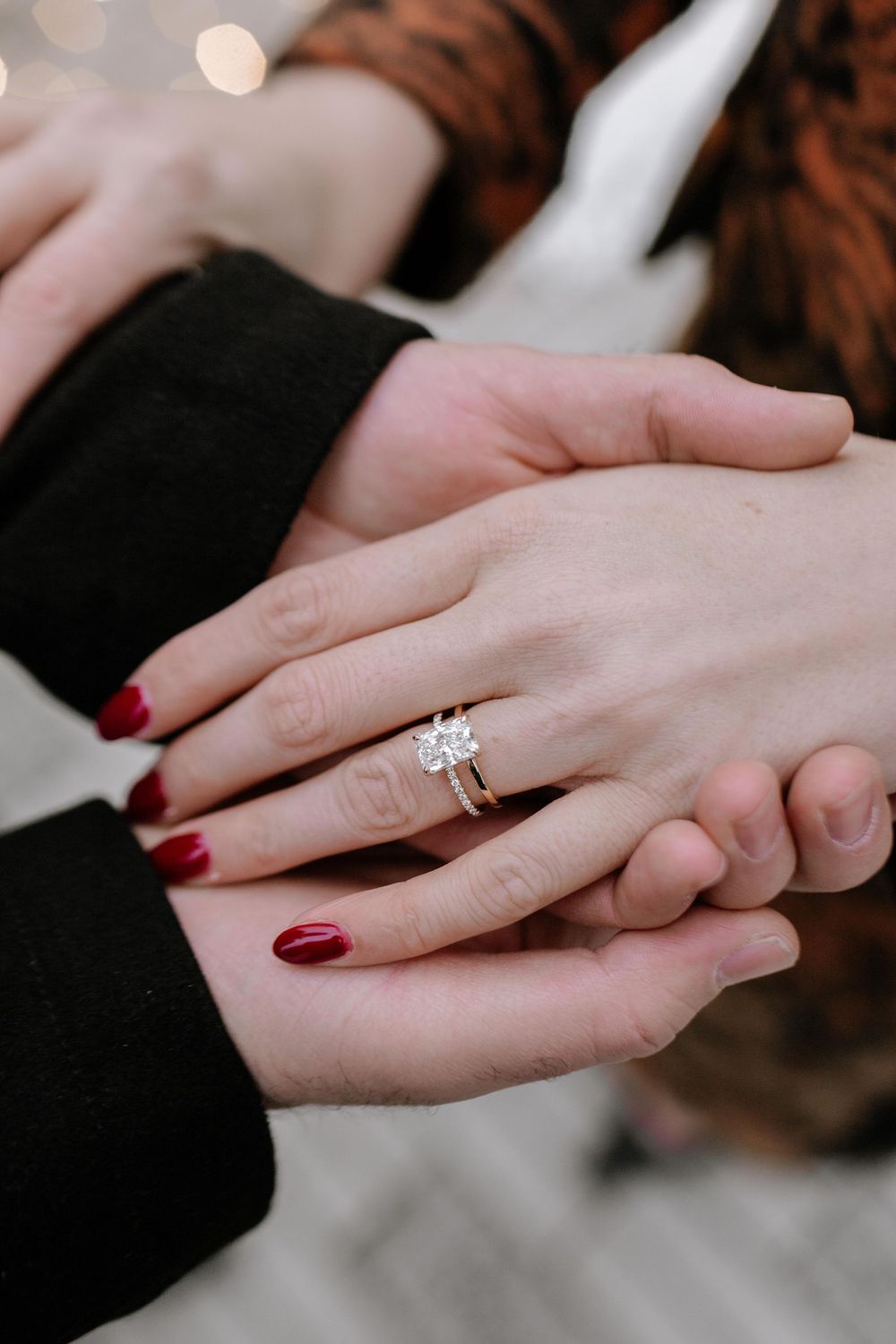 Close-up of hands exchanging engagement ring with red nail polish against dark background.