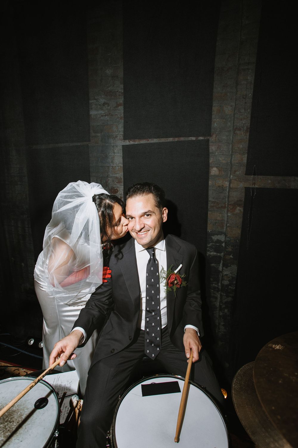 Groom in black suit plays drums with drumsticks while bride in white veil kisses his cheek at wedding reception.