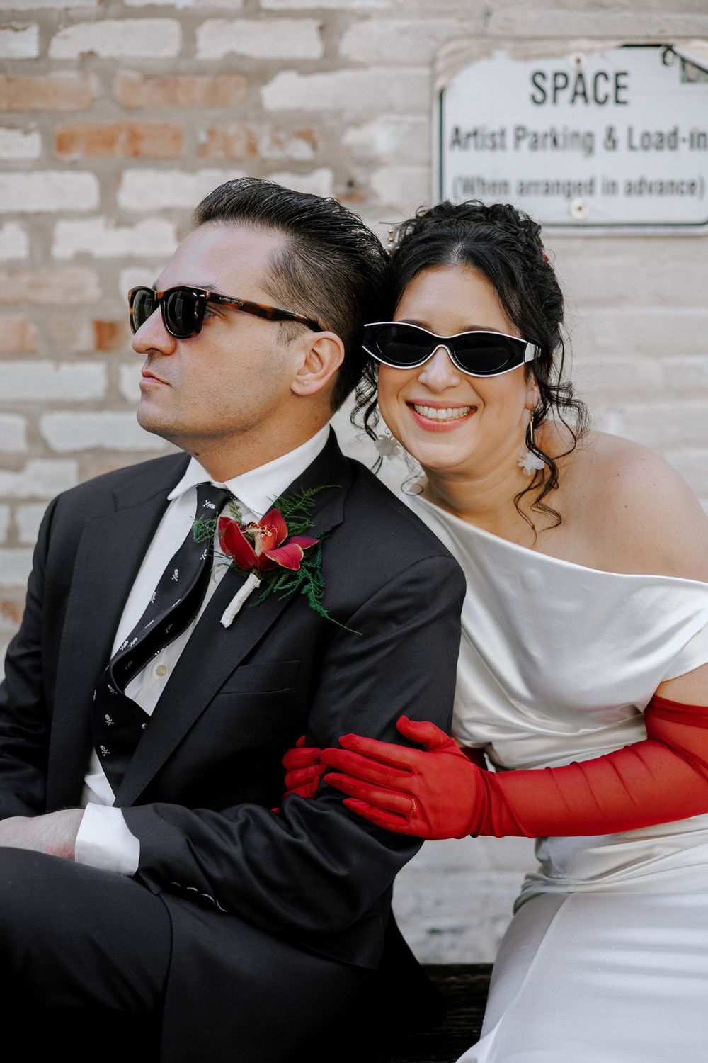Stylish bride and groom wearing matching black sunglasses sitting together outdoors against brick wall at wedding.