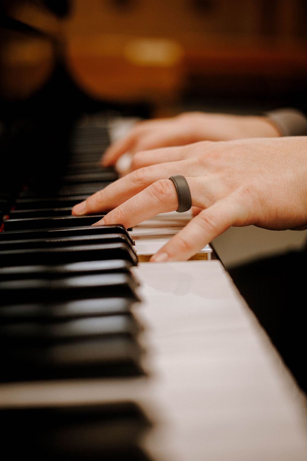 Close-up of hands playing piano keys, one wearing a dark ring, with blurred guitar in background.