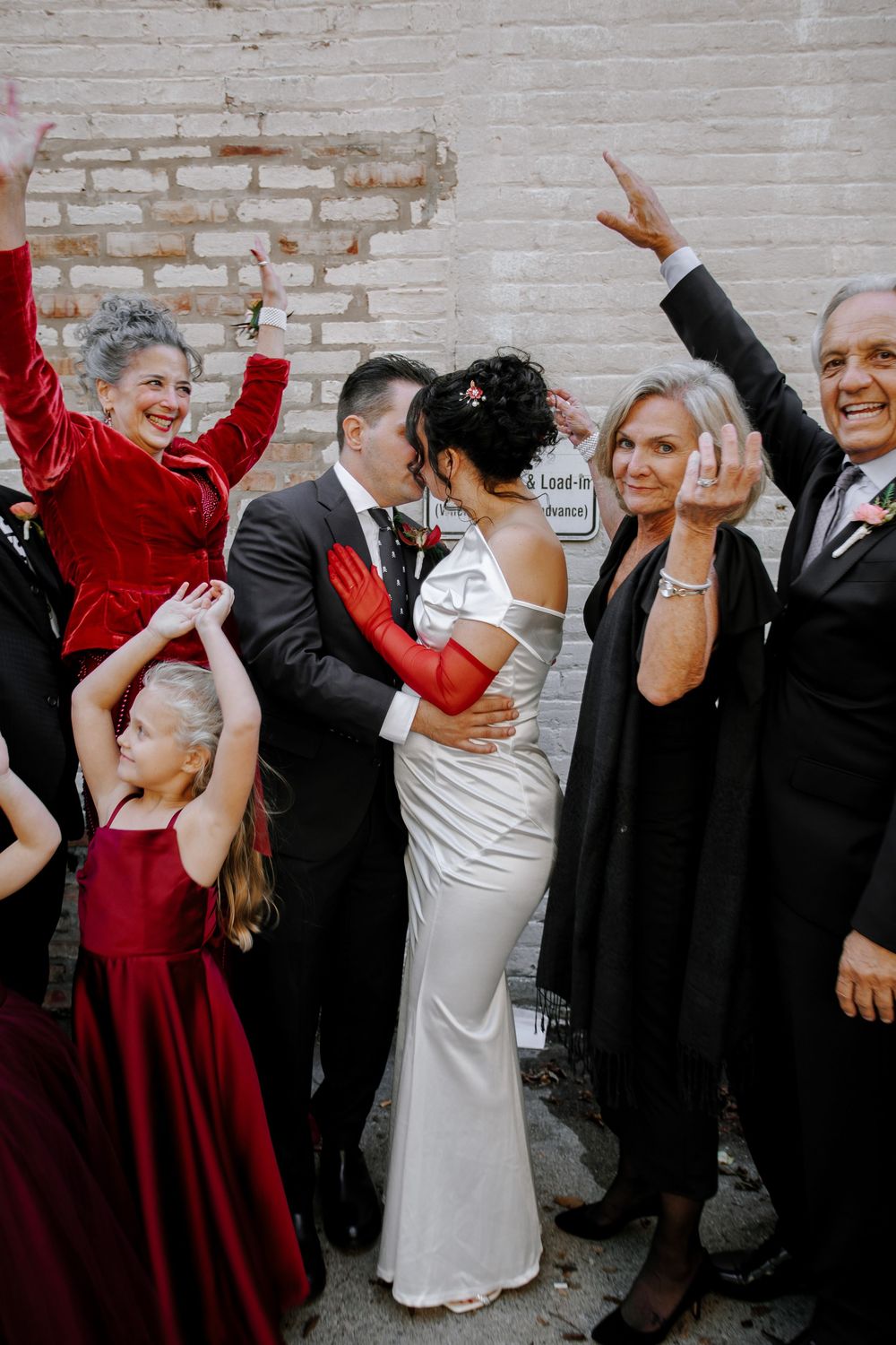 Joyful wedding couple kisses surrounded by cheering family members in red dresses outside a brick building.