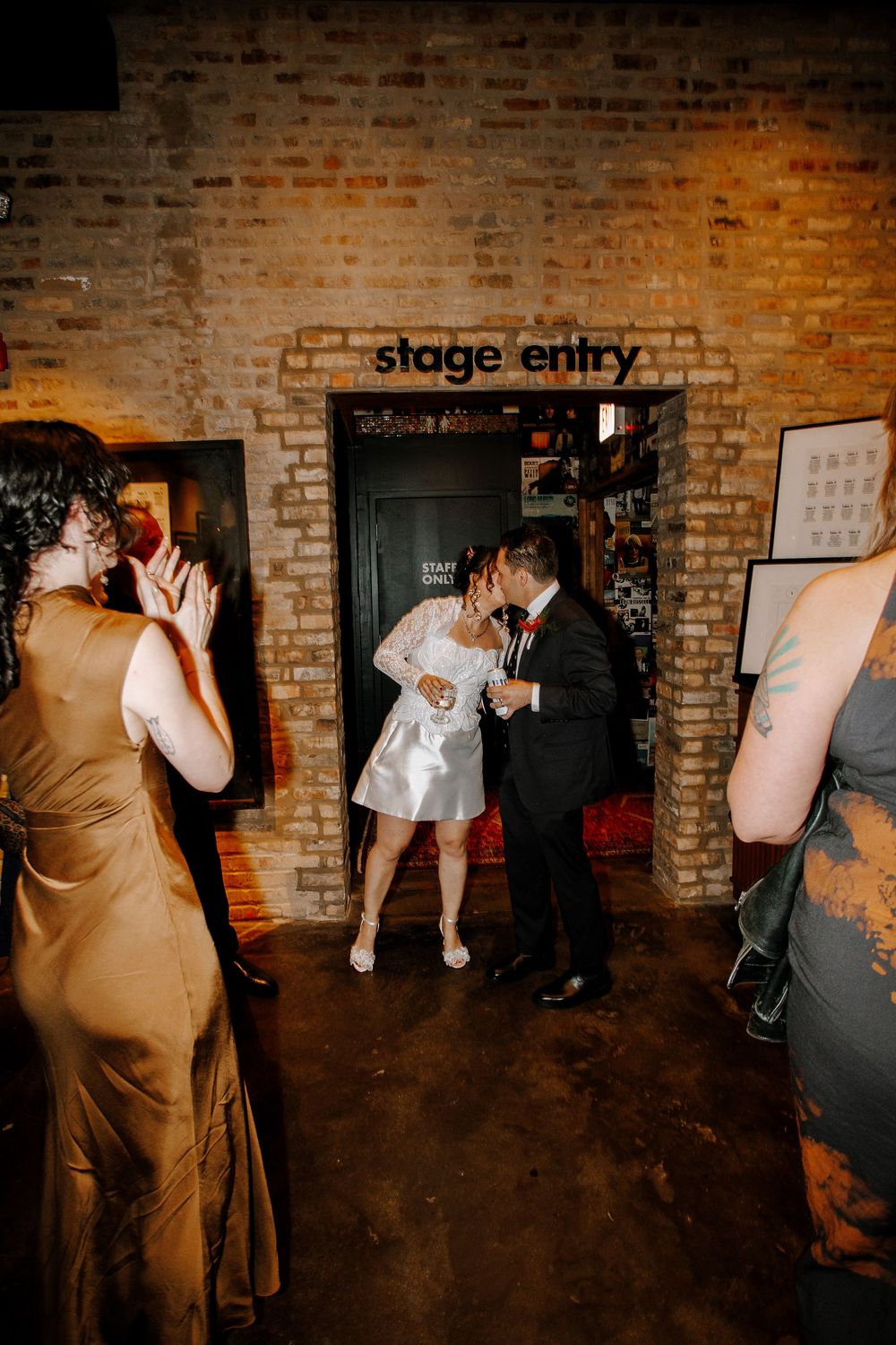 Bride and groom share a kiss at stage entry doorway while wedding guests applaud in brick venue.
