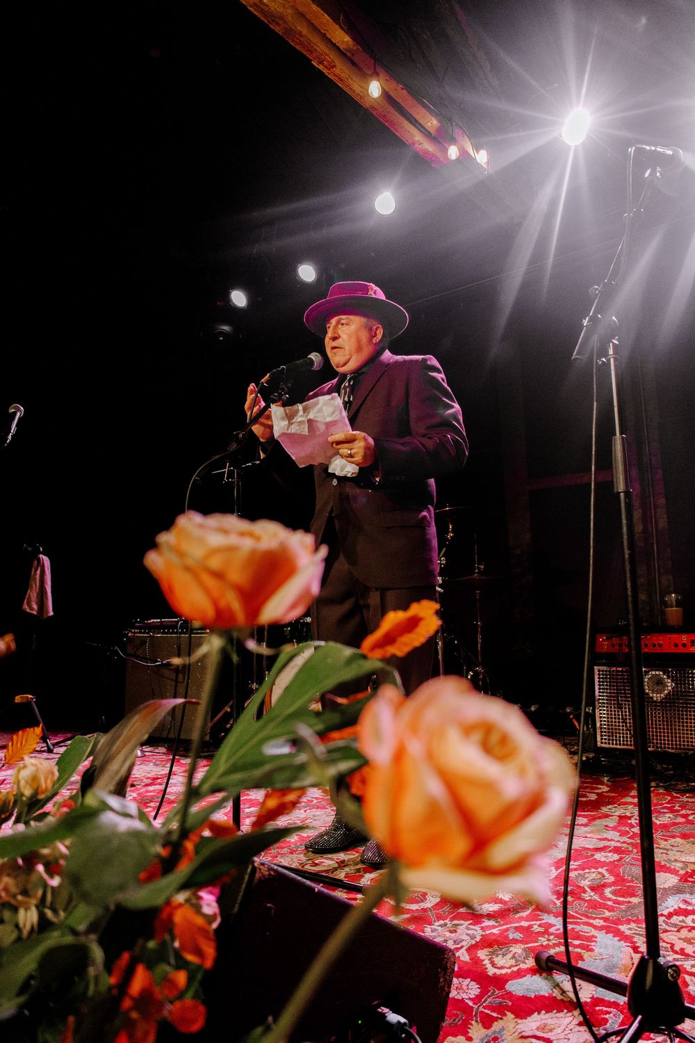 Elderly man in purple hat reading at microphone on stage with orange roses in foreground.