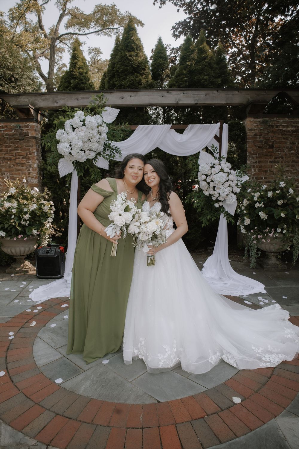 Wedding party in olive green dresses poses with white floral arrangements on a brick patio.