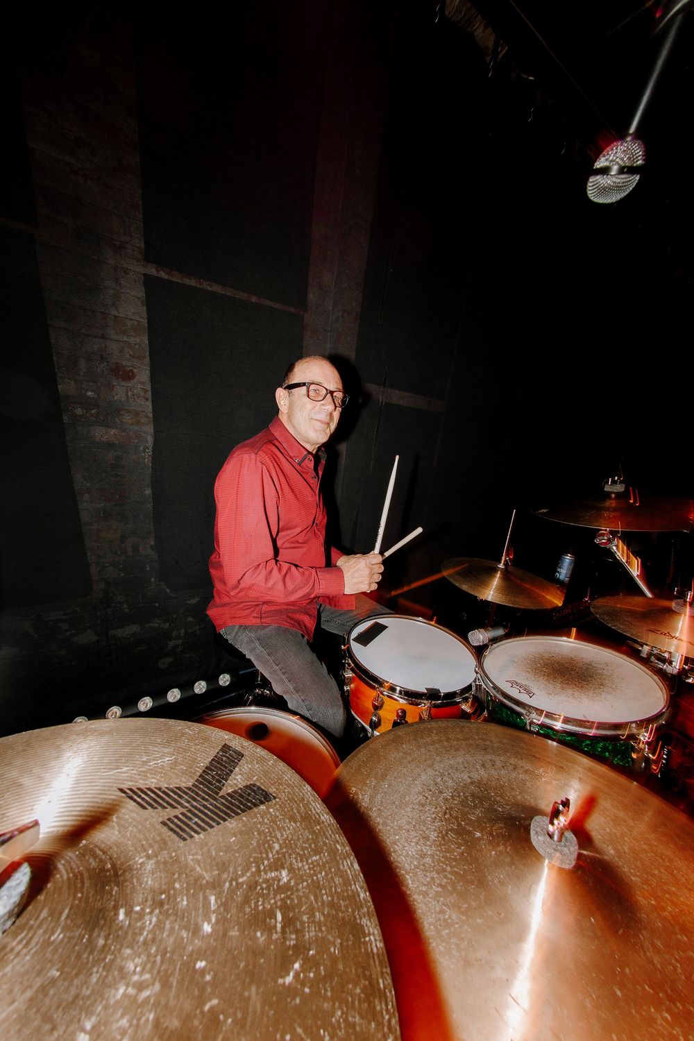 Smiling drummer in red shirt plays drum kit with drumsticks in dimly lit concert venue stage.