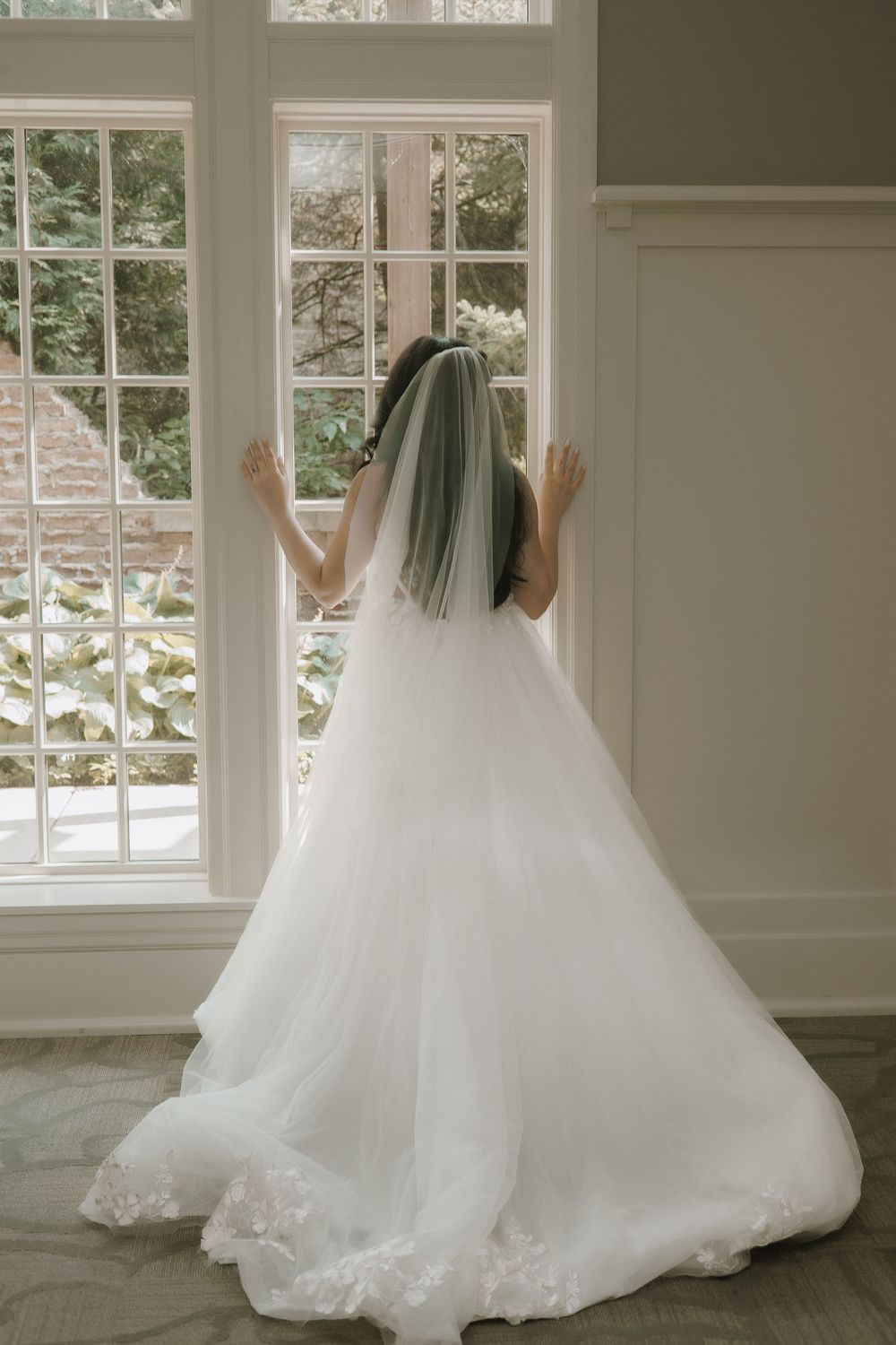 A bride in a flowing white wedding gown stands near large windows in an elegant room, creating a romantic silhouette.