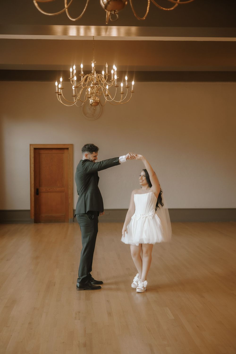 A couple dances together in a spacious ballroom with hardwood floors and elegant chandeliers overhead.