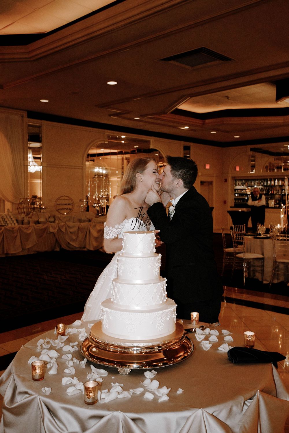 Bride and groom kissing behind elegant four-tier white wedding cake at reception venue.