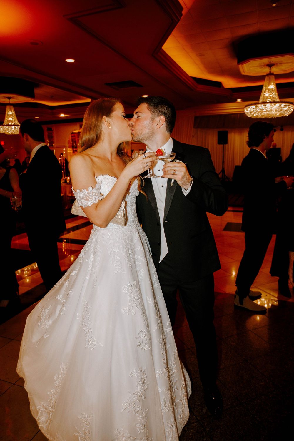 Bride and groom share a kiss on the dance floor while holding martini glasses at their wedding reception.