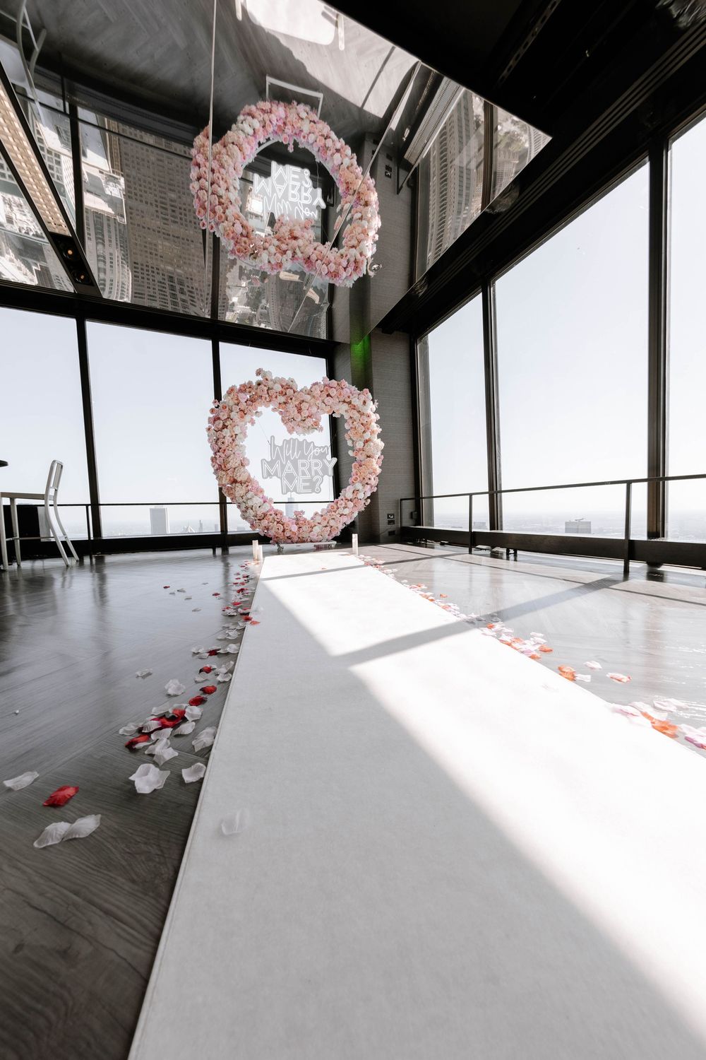 Pink floral arches line a sunlit indoor space with floor rose petals creating an elegant proposal setting.