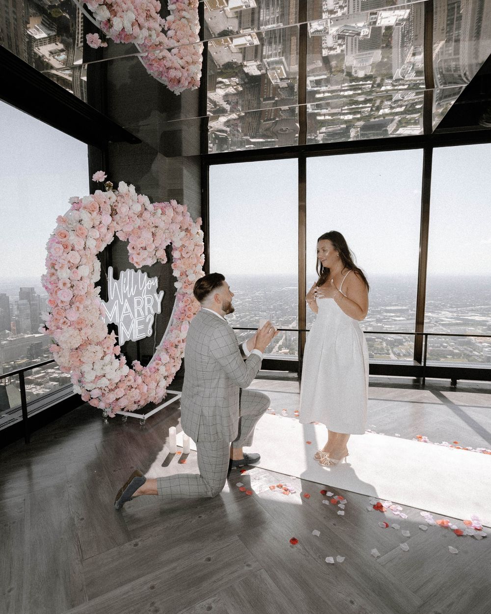 A romantic proposal scene unfolds in a modern high-rise space with pink cherry blossom decorations and city views.