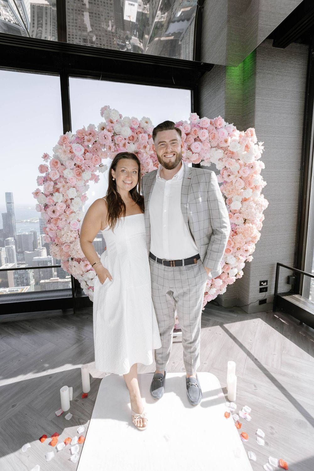 Couple poses together in front of pink balloon arch in modern indoor space with city views.