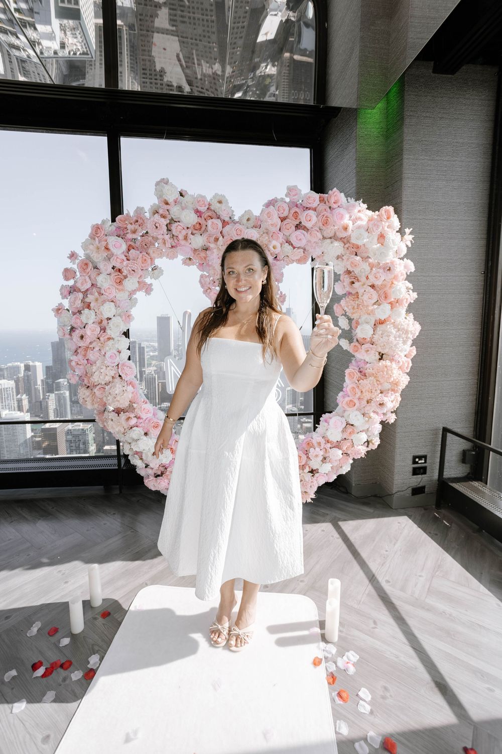 A heart-shaped pink floral arch installation frames a modern interior space with city views through large windows.