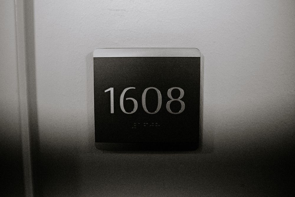 Black hotel room number sign displaying 1608 with Braille text mounted on a white wall.