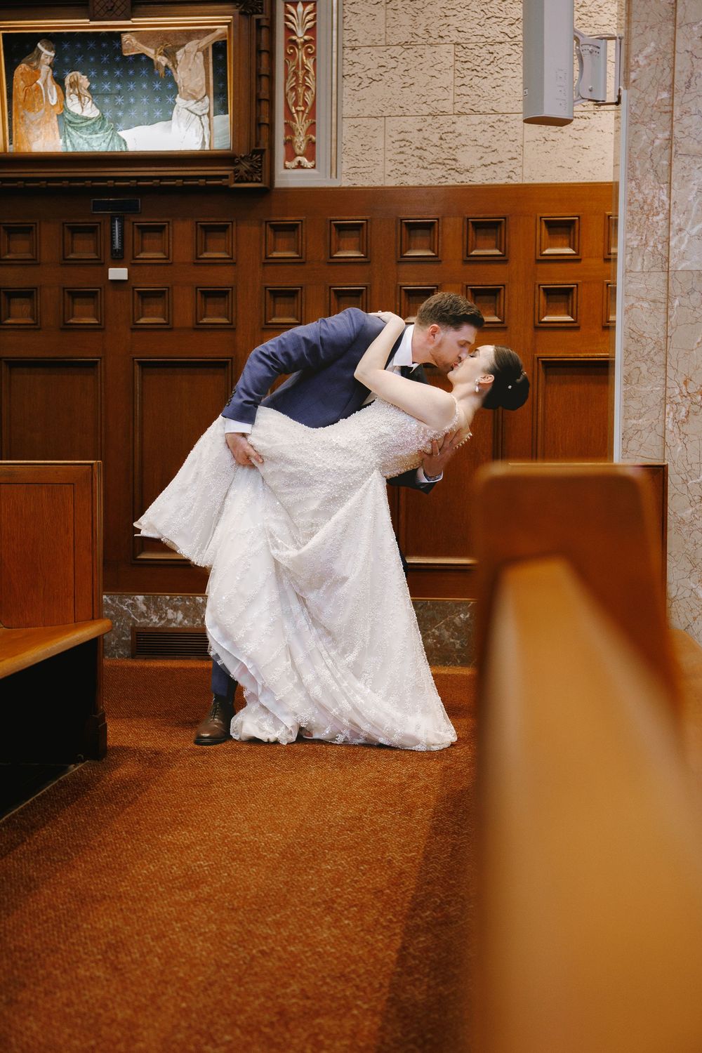 Groom dips bride for a romantic kiss in a church aisle, bride in white lace gown, groom in navy suit.