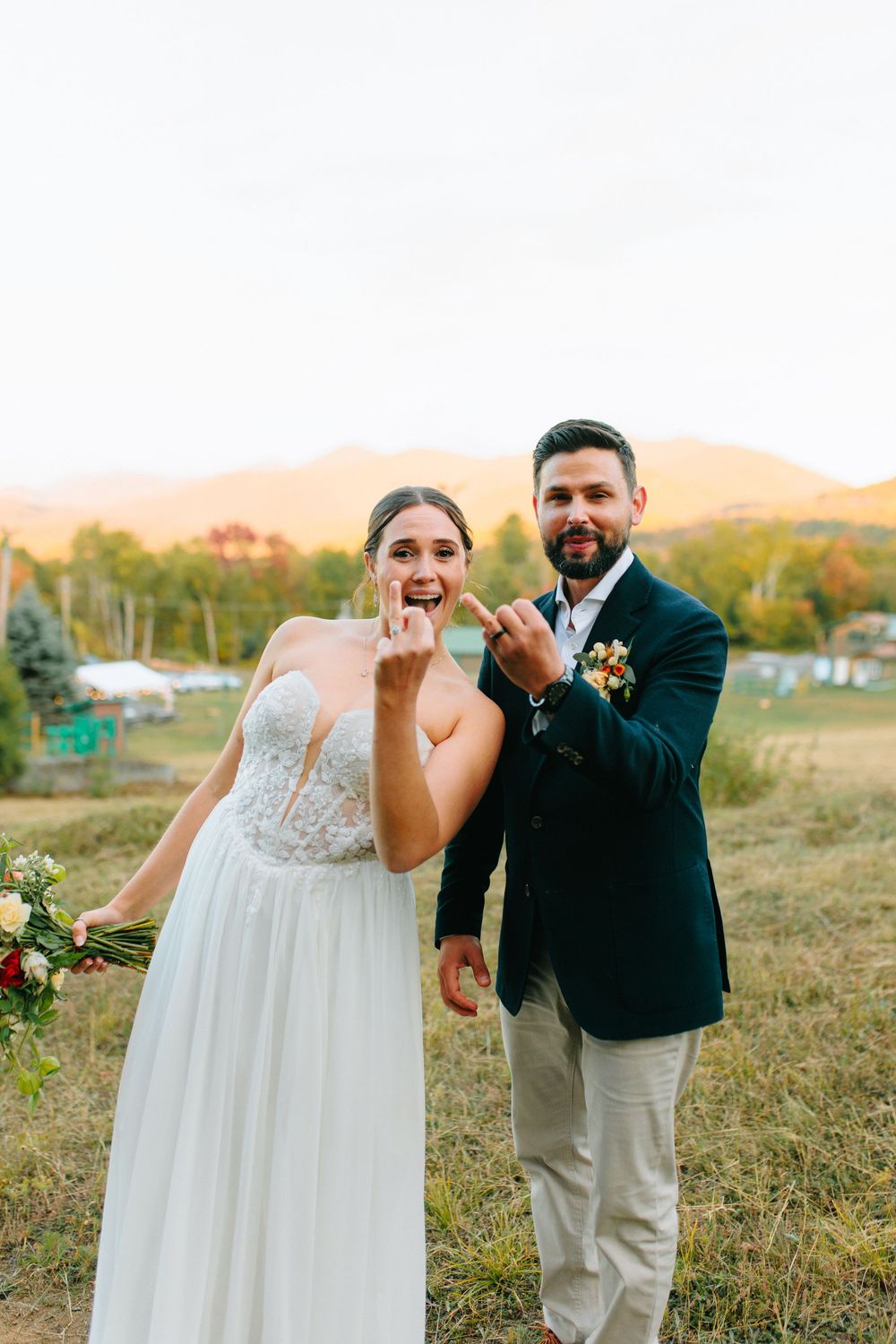 A joyful wedding couple showing off their rings in an outdoor setting with mountains in the background at sunset.
