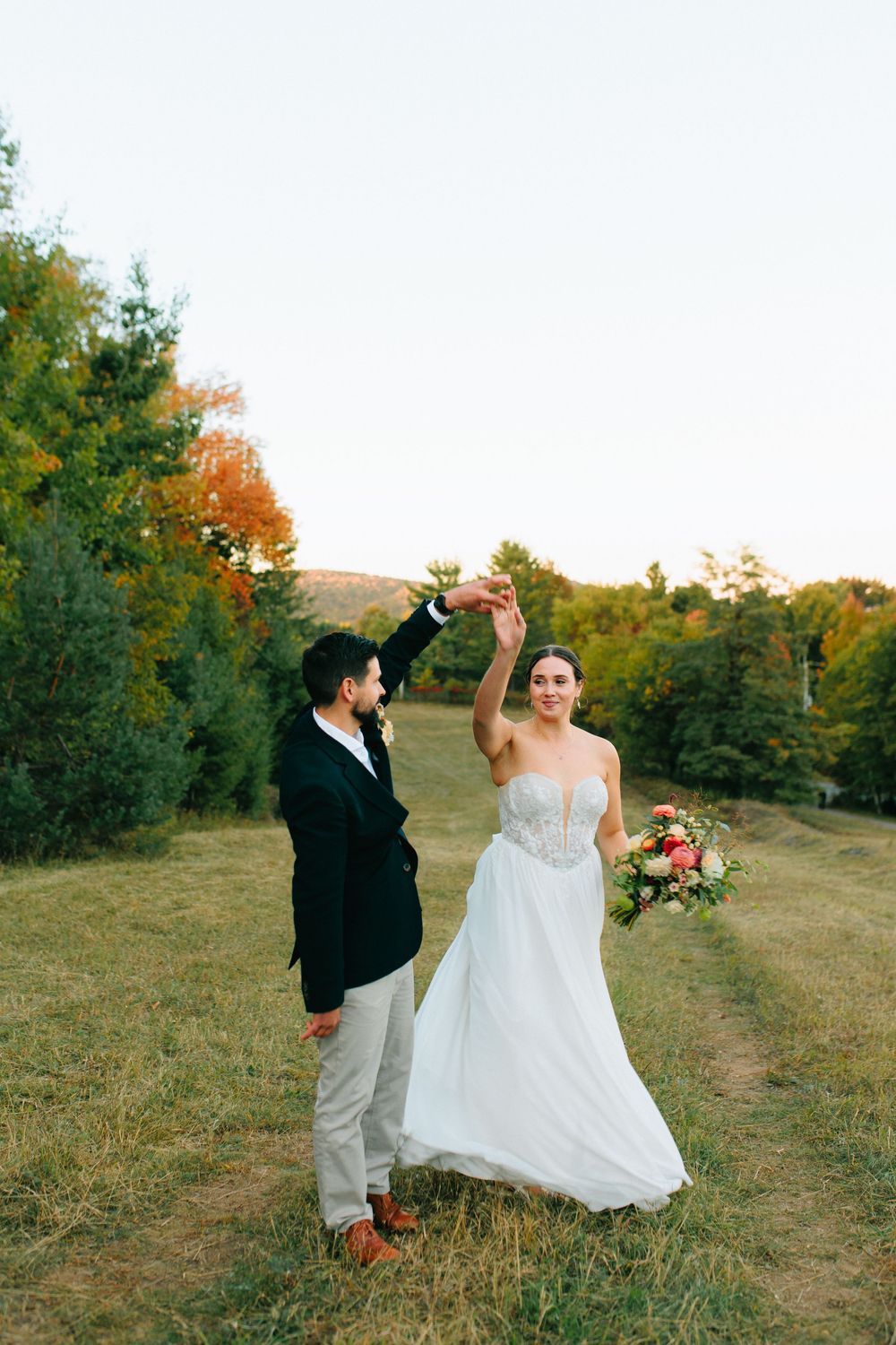 A couple dances together in a field during golden hour with autumn trees in the background.